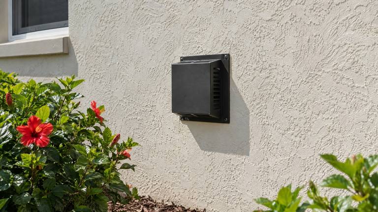 A black direct-vent termination cap on a cream stucco exterior wall above a hibiscus bed.