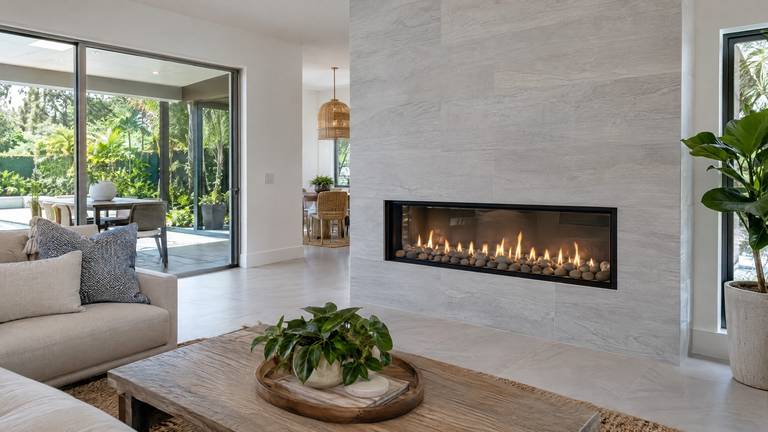A new linear gas fireplace with a row of flames over river rock in a light gray tile wall.